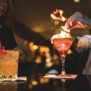 Close-up of a pink cocktail lit by flames being placed on a table