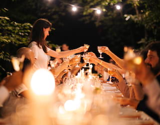 Cut crystal wine glasses shine in candlelight as they are held aloft in a toast by a long table of diners in a night garden