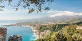 View from Capo Taormina south over the Ionian coast, with pretty seaside towns hemmed in by bright sea and steep green hills.