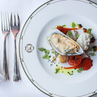 Looking down on a branded Eastern and Oriental Express dining plate with a cooked mussel starter presented with garnish.