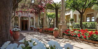 Bougainvillea drapes from an iron pergola shading a large doorway. The entrance is lined by red geraniums and white petunias