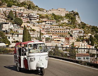 A classic white Piaggeo Ape offers guests great views beyond its open red cabriolet roof, as it explores Taormina's streets