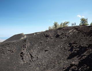Tourists standing on the dark, rocky slopes of Mount Etna under blue skies
