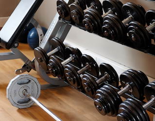 Fitness centre detail of a weights rack with rows of black dumbbells and a long silver weighted barbell on the floor.