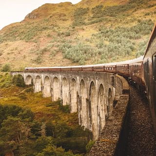 Looking from a window to the front of the train, which pulls carriages over the iconic Glenfinnan Viaduct towards a hillside.