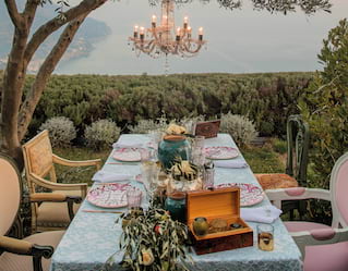 The garden table is set for a celebration beneath a chandelier suspended from an olive tree, with breathtaking coastal views.